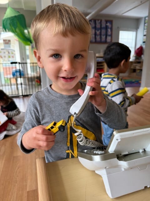 Pre‑K student using toy phone and cash register to explore technology concepts through imaginative role play at Kids Konnect